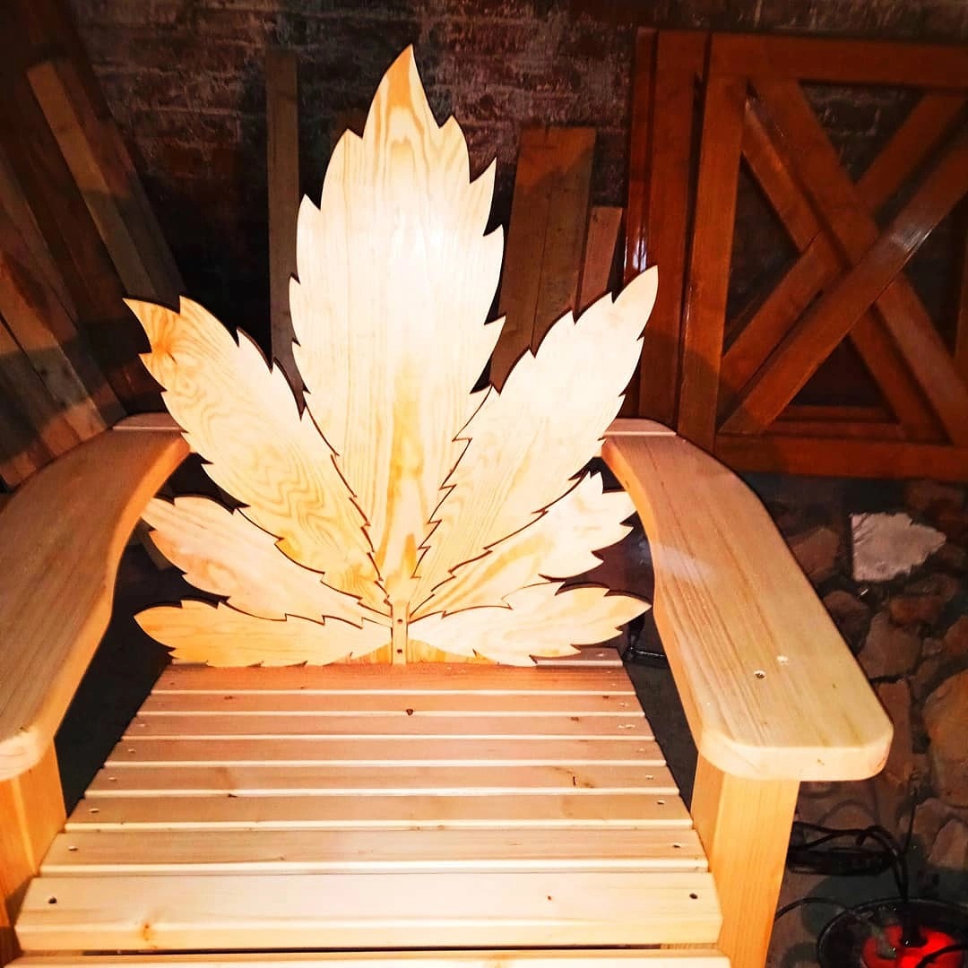 Drewniany fotel z oparciem w kształcie liścia klonu, wykonany z jasnego drewna, widoczne słoje, na tle ceglanej ściany i drewnianych elementów konstrukcyjnych.