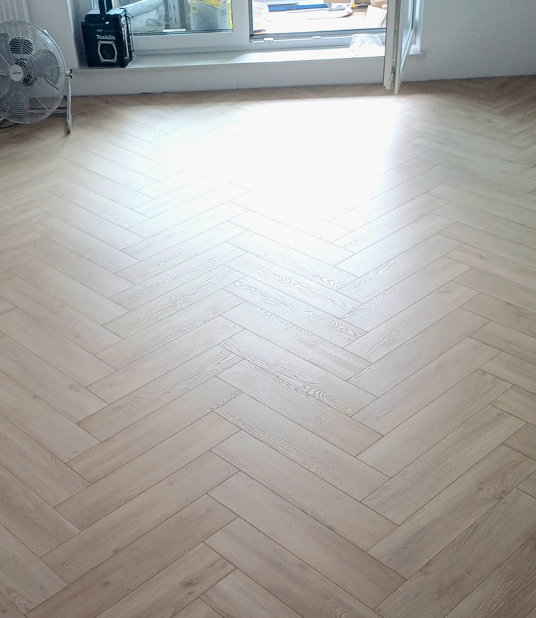 Jasne panele podłogowe ułożone w jodełkę francuską, widoczny wentylator i głośnik Makita przy oknie.