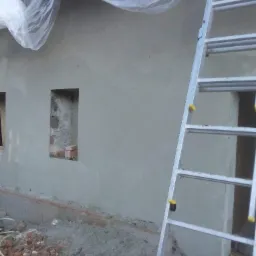 Świeżo otynkowana ściana zewnętrzna z otworami okiennymi, zabezpieczona folią od góry, z aluminiową drabiną opartą o ścianę i gruzem ceglanym u podstawy.