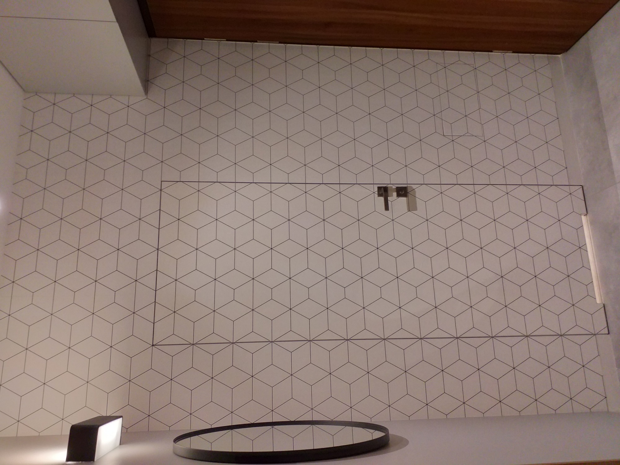 Ściana w łazience wykończona białymi płytkami z szarym geometrycznym wzorem w kształcie sześcianów, z widoczną ukrytą szafką i elementami armatury.