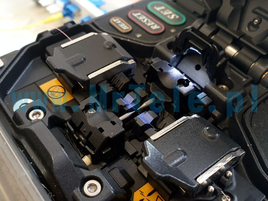 Zbliżenie na spawarkę światłowodową podczas łączenia włókien, widoczne uchwyty, elektrody i włókno światłowodowe.