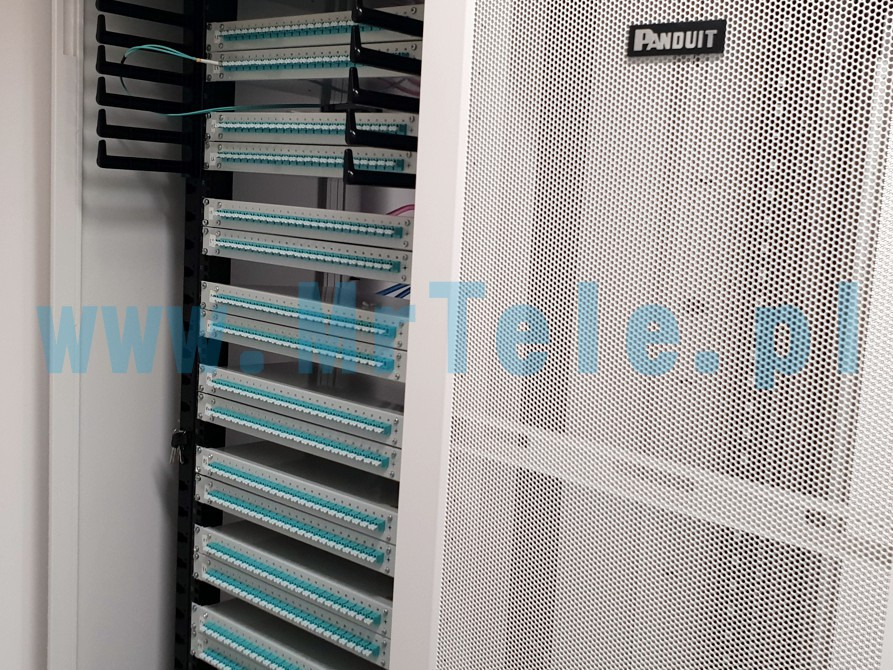 Panel krosowniczy Panduit w szafie rack, widoczne porty światłowodowe z niebieskimi wkładkami, czarne uchwyty na kable po lewej stronie.