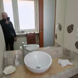 Okrągła umywalka nablatowa z chromowaną baterią na blacie wykończonym mozaiką; w tle lustro odbijające fragment łazienki i mężczyznę robiącego zdjęcie.