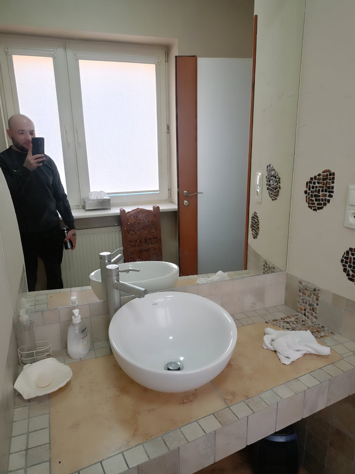 Okrągła umywalka nablatowa z chromowaną baterią na blacie wykończonym mozaiką; w tle lustro odbijające fragment łazienki i mężczyznę robiącego zdjęcie.