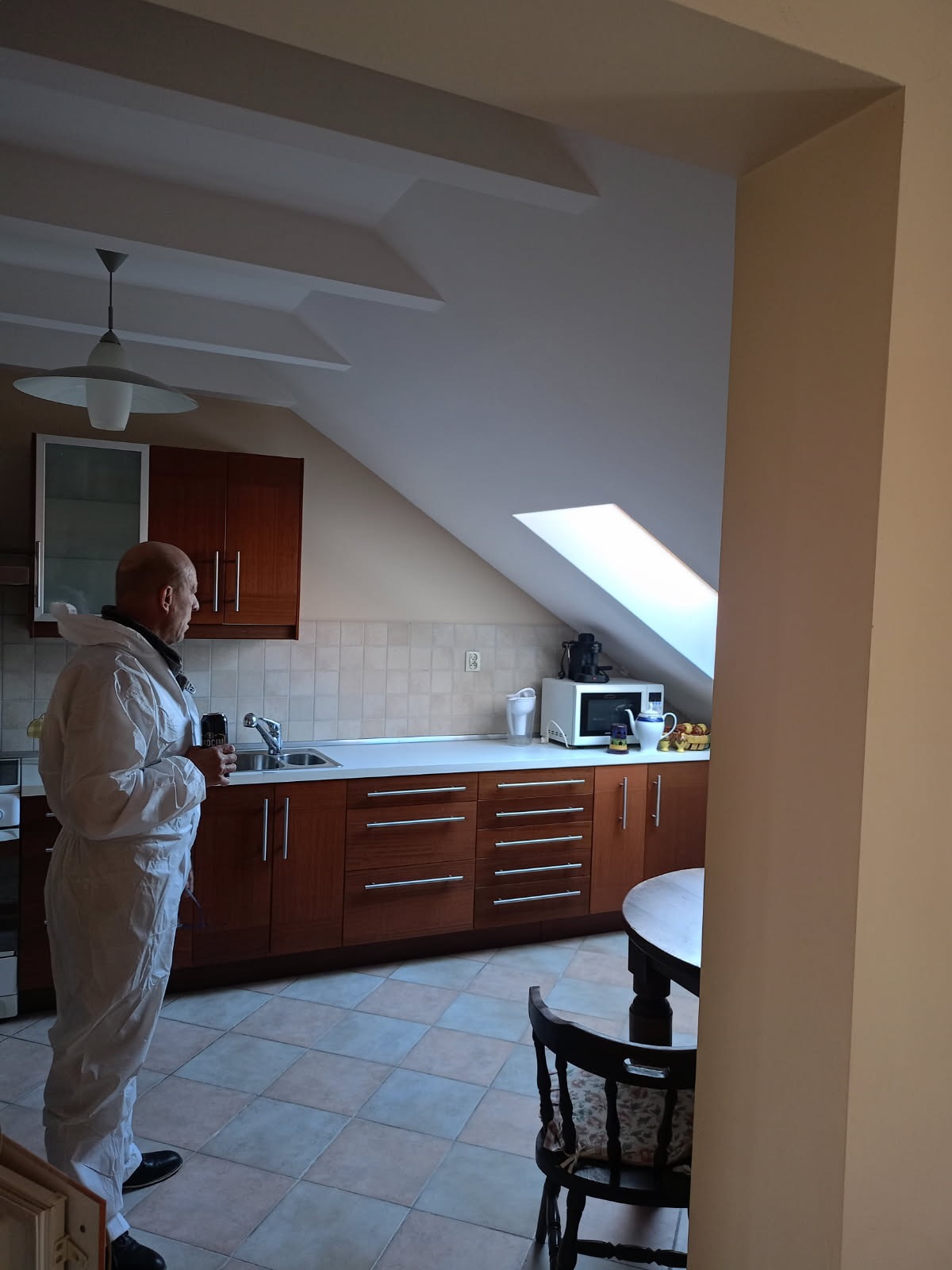 Pracownik w kombinezonie ochronnym w kuchni z oknem dachowym, przed szafkami z ciemnego drewna i jasnym blatem, płytki podłogowe w szaro-beżową kratę.