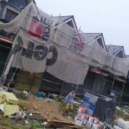 GRAC-BUD - Dom w trakcie prac elewacyjnych, rusztowania osłonięte siatką z nadrukiem reklamowym, widoczny pracownik i materiały budowlane.