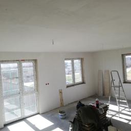 GRAC-BUD - Pokój w trakcie remontu: świeżo otynkowane ściany i sufit, okna z folią ochronną, drabina aluminiowa, wiadro z farbą i sterta płyt na podłodze.