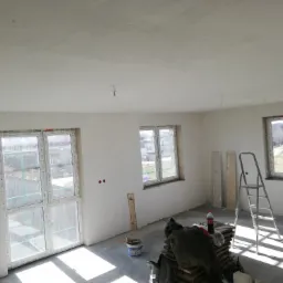 Pokój w trakcie remontu: świeżo otynkowane ściany i sufit, okna z folią ochronną, drabina aluminiowa, wiadro z farbą i sterta płyt na podłodze.