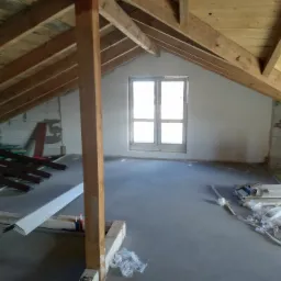 Poddasze w trakcie adaptacji: widoczne drewniane krokwie, surowa wylewka, okno dachowe, materiały budowlane i narzędzia przygotowane do dalszych prac wykończeniowych.