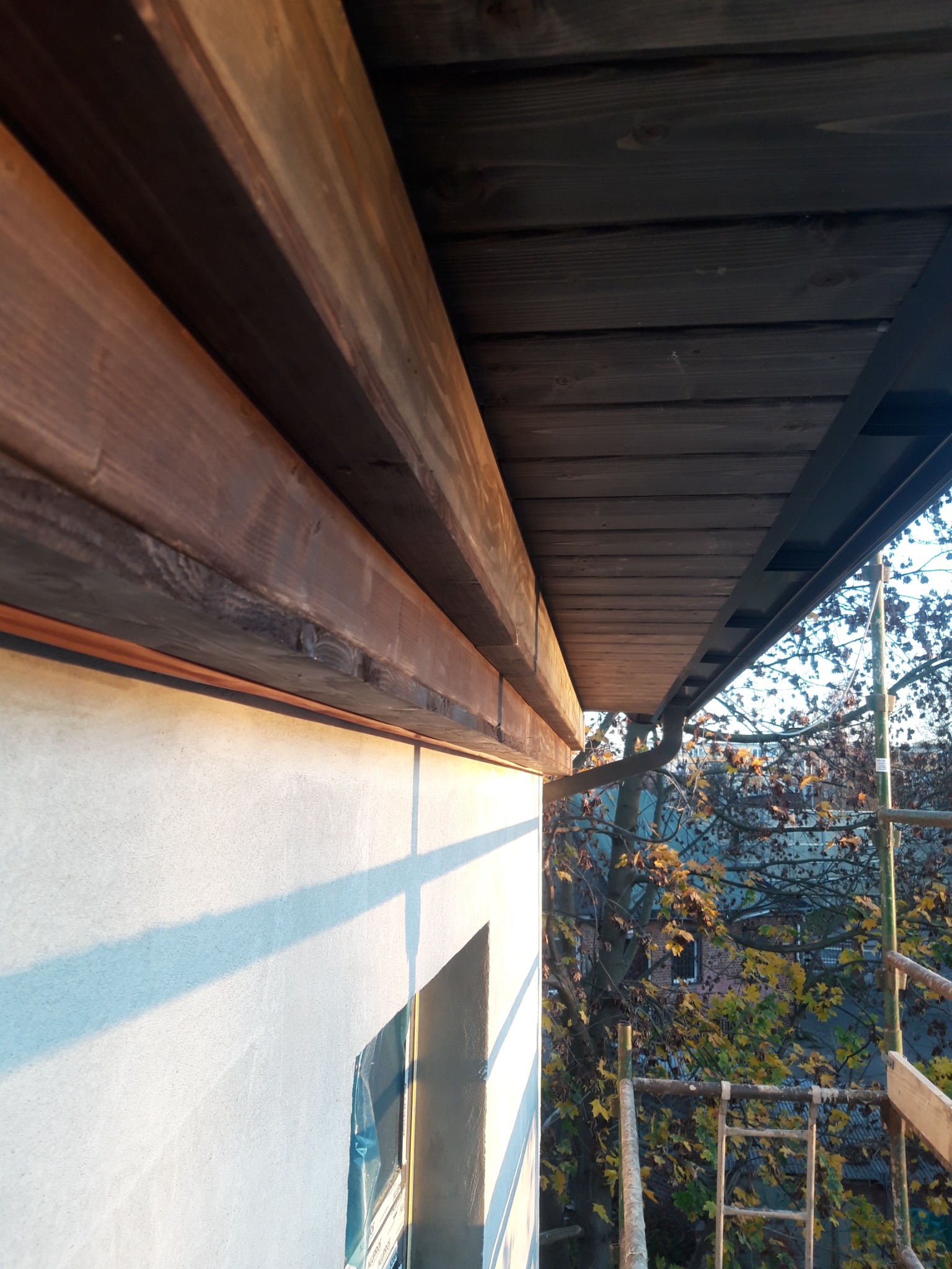 Drewniana podbitka dachowa z widocznymi elementami konstrukcyjnymi, elewacja budynku w trakcie prac wykończeniowych, rusztowanie i jesienne drzewo w tle.