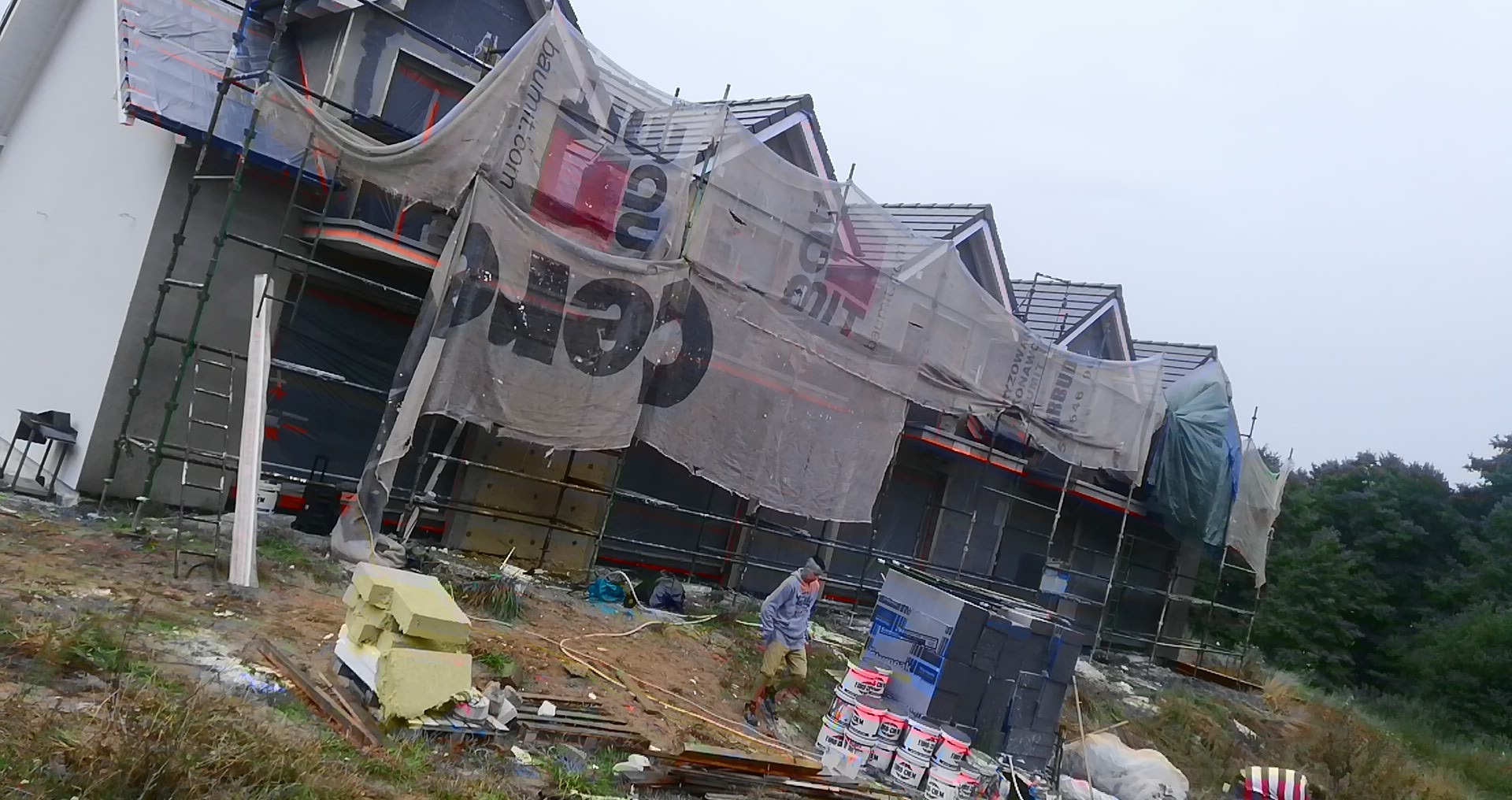 Dom w trakcie prac elewacyjnych, rusztowania osłonięte siatką z nadrukiem reklamowym, widoczny pracownik i materiały budowlane.