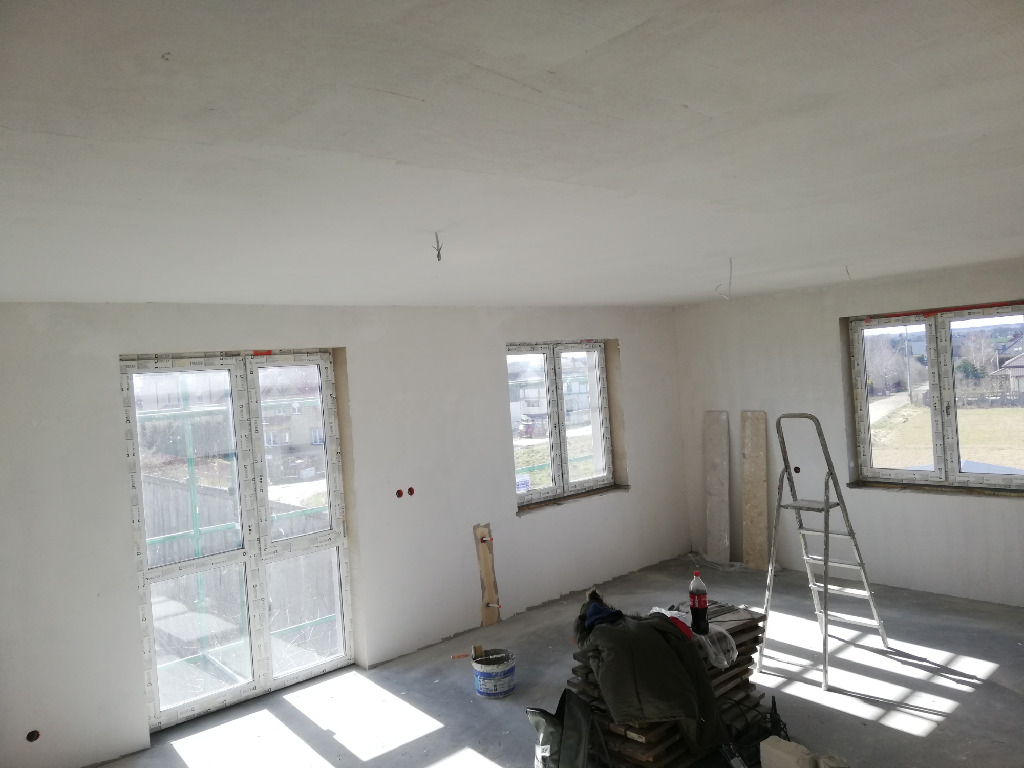 Pokój w trakcie remontu: świeżo otynkowane ściany i sufit, okna z folią ochronną, drabina aluminiowa, wiadro z farbą i sterta płyt na podłodze.