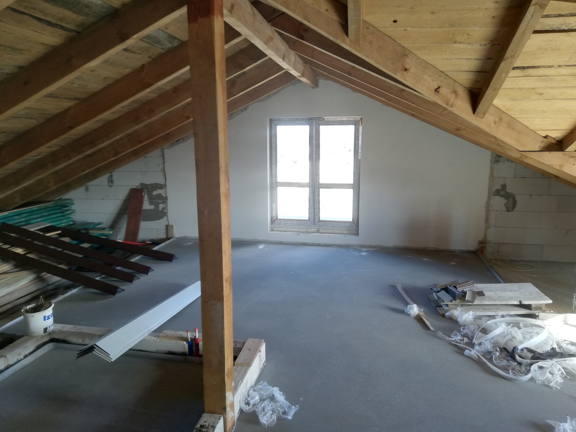 Poddasze w trakcie adaptacji: widoczne drewniane krokwie, surowa wylewka, okno dachowe, materiały budowlane i narzędzia przygotowane do dalszych prac wykończeniowych.