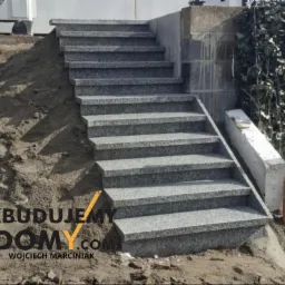 Granitowe schody zewnętrzne z szarego kamienia, prowadzące w górę zbocza, z widocznym fragmentem budynku i roślinnością w tle. Detal wykończenia ogrodu.