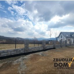 zbudujemydomy.com - Betonowo-metalowe ogrodzenie panelowe z podmurówką, widok z zewnątrz na tle domu i pagórkowatego krajobrazu pod pochmurnym niebem.