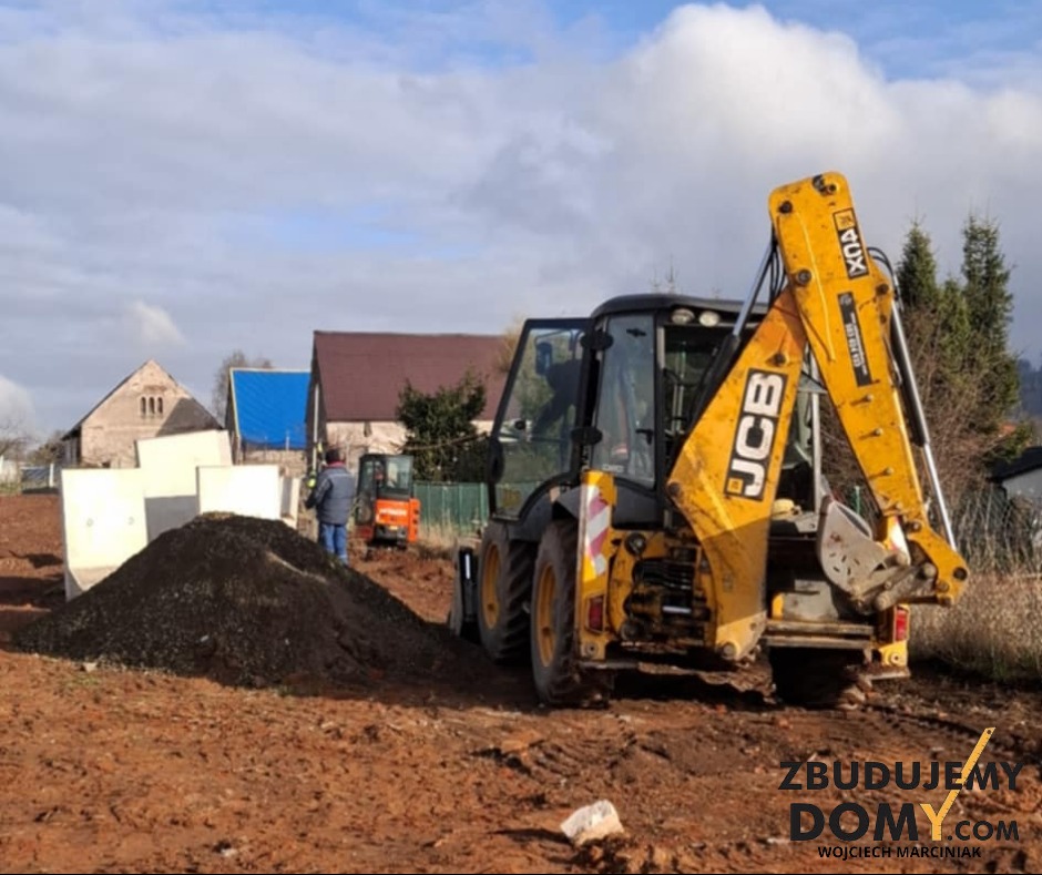 Żółta koparko-ładowarka JCB na placu budowy z przygotowanym wykopem pod fundamenty, w tle budynki mieszkalne i pochmurne niebo.