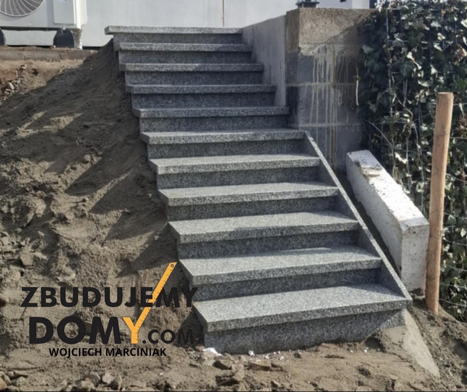 Granitowe schody zewnętrzne z szarego kamienia, prowadzące w górę zbocza, z widocznym fragmentem budynku i roślinnością w tle. Detal wykończenia ogrodu.