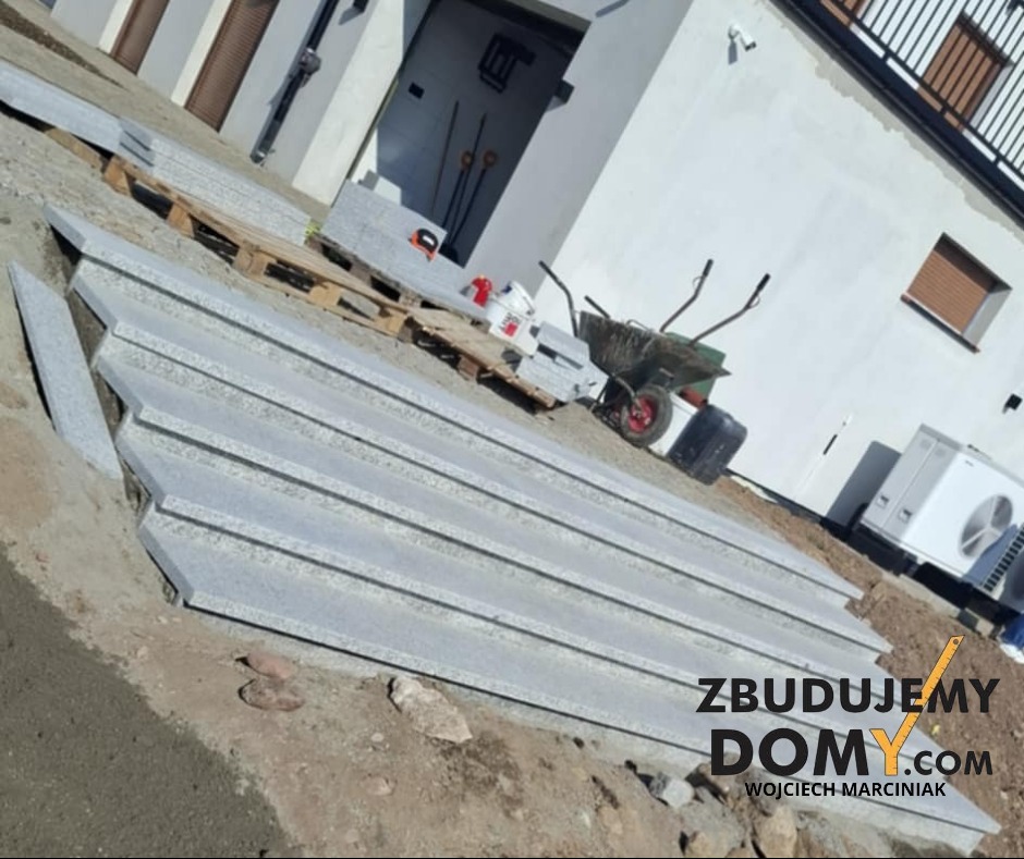 Granitowe stopnie schodowe ułożone na ziemi przed budynkiem, przygotowane do montażu. Widoczna taczka i materiały budowlane w tle. Ukośne ujęcie.