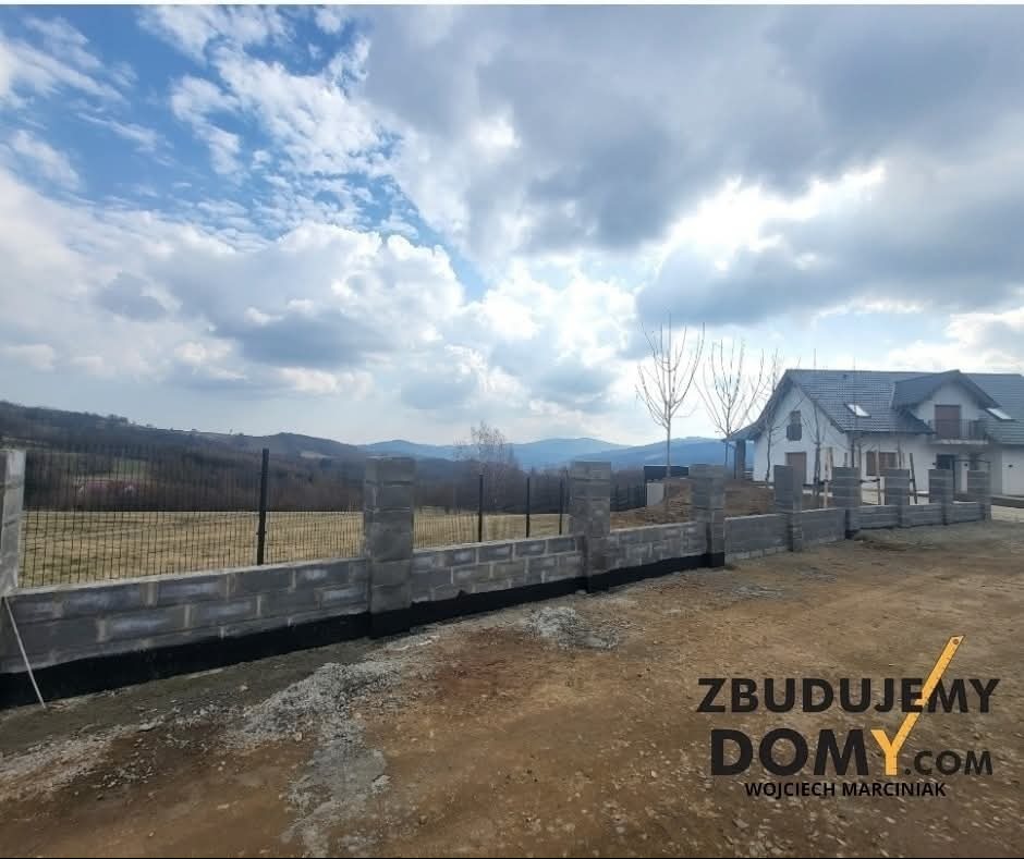 Betonowo-metalowe ogrodzenie panelowe z podmurówką, widok z zewnątrz na tle domu i pagórkowatego krajobrazu pod pochmurnym niebem.