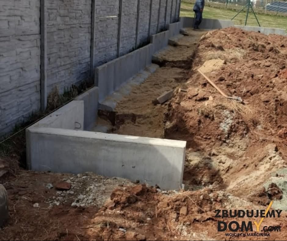 Betonowy mur oporowy w kształcie litery L w trakcie budowy, zabezpieczający teren przed osuwaniem. Widoczny wykop i częściowo zasypany piaskiem. W tle ogrodzenie panelowe.