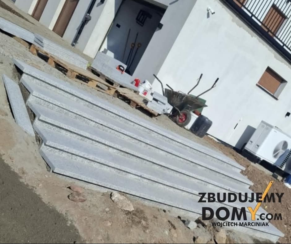 Granitowe stopnie schodowe przed budynkiem, przygotowane do montażu. W tle taczka, paleta i fragment budynku z klimatyzatorem. Widoczne logo firmy.