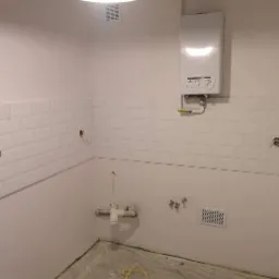 Ściana w trakcie układania białych, prostokątnych płytek, widoczne otwory na gniazdka i rury, z zamontowanym piecykiem gazowym i kratką wentylacyjną, podłoga zabezpieczona folią.