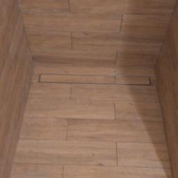 Niemakoś - Wykończona kabina prysznicowa z odpływem liniowym, wyłożona płytkami imitującymi drewno, widoczne kartony i akcesoria montażowe na podłodze.