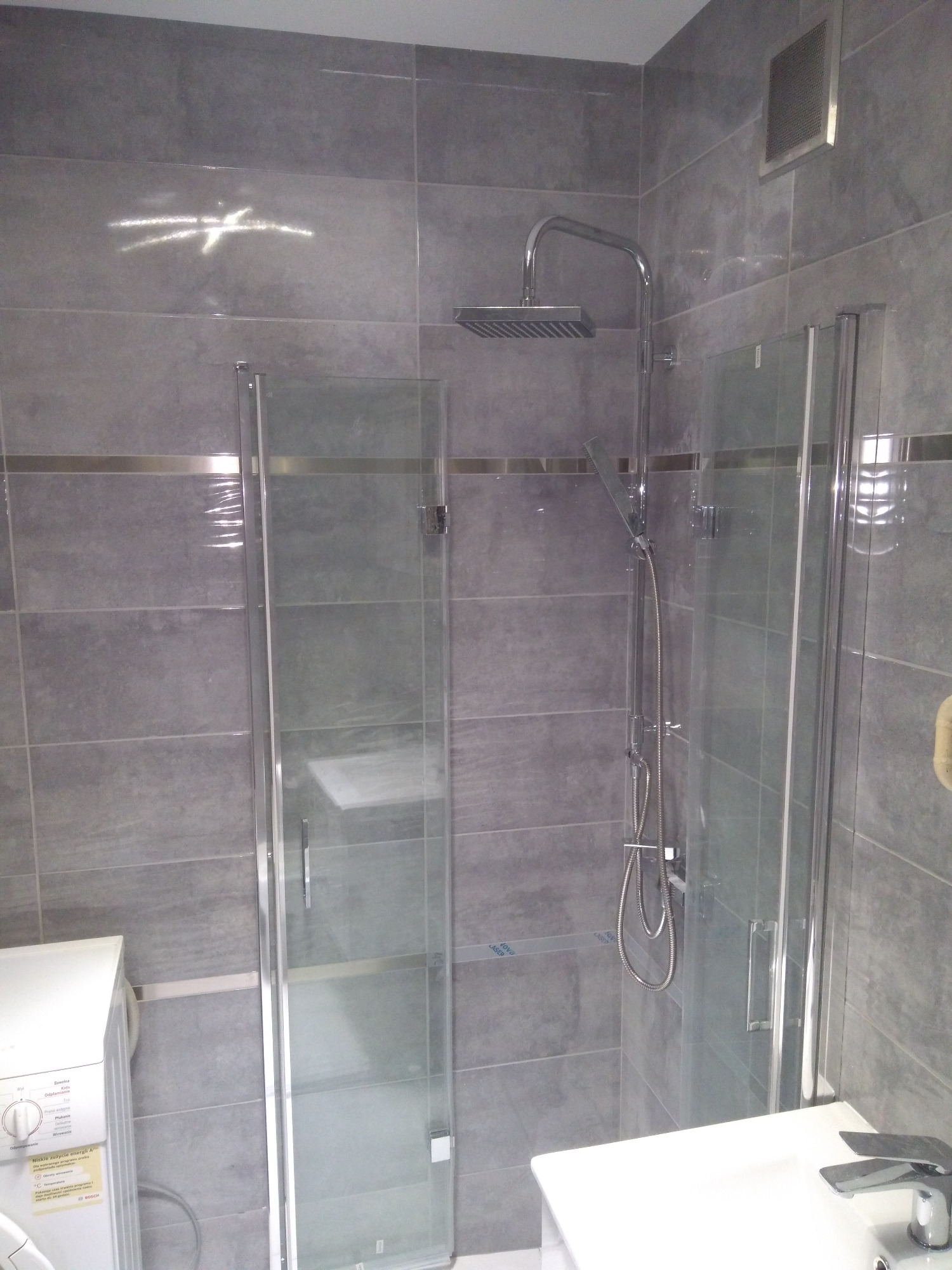 Nowoczesna łazienka z szarymi płytkami, kabiną prysznicową ze szklanymi drzwiami, deszczownicą i baterią prysznicową, widoczna pralka z naklejką informacyjną.