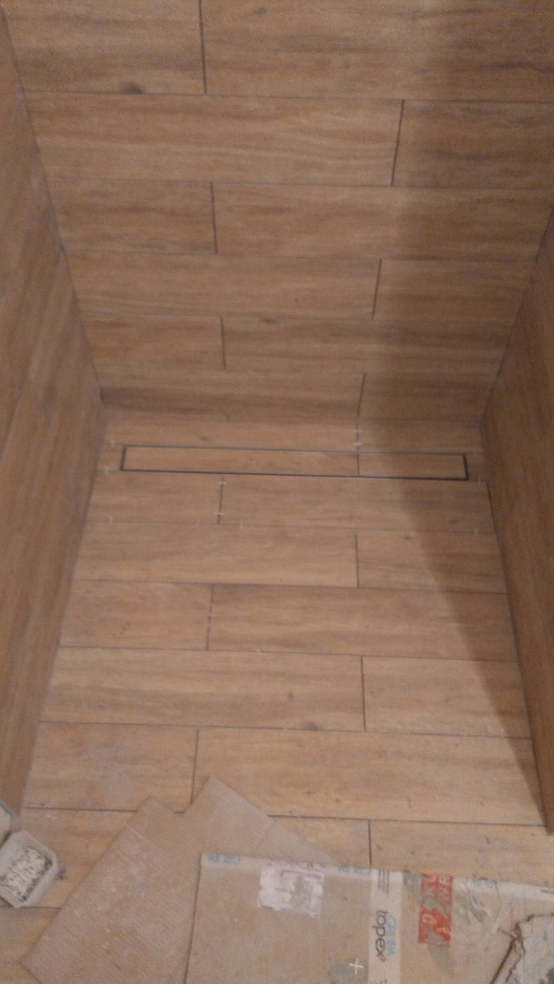 Wykończona kabina prysznicowa z odpływem liniowym, wyłożona płytkami imitującymi drewno, widoczne kartony i akcesoria montażowe na podłodze.