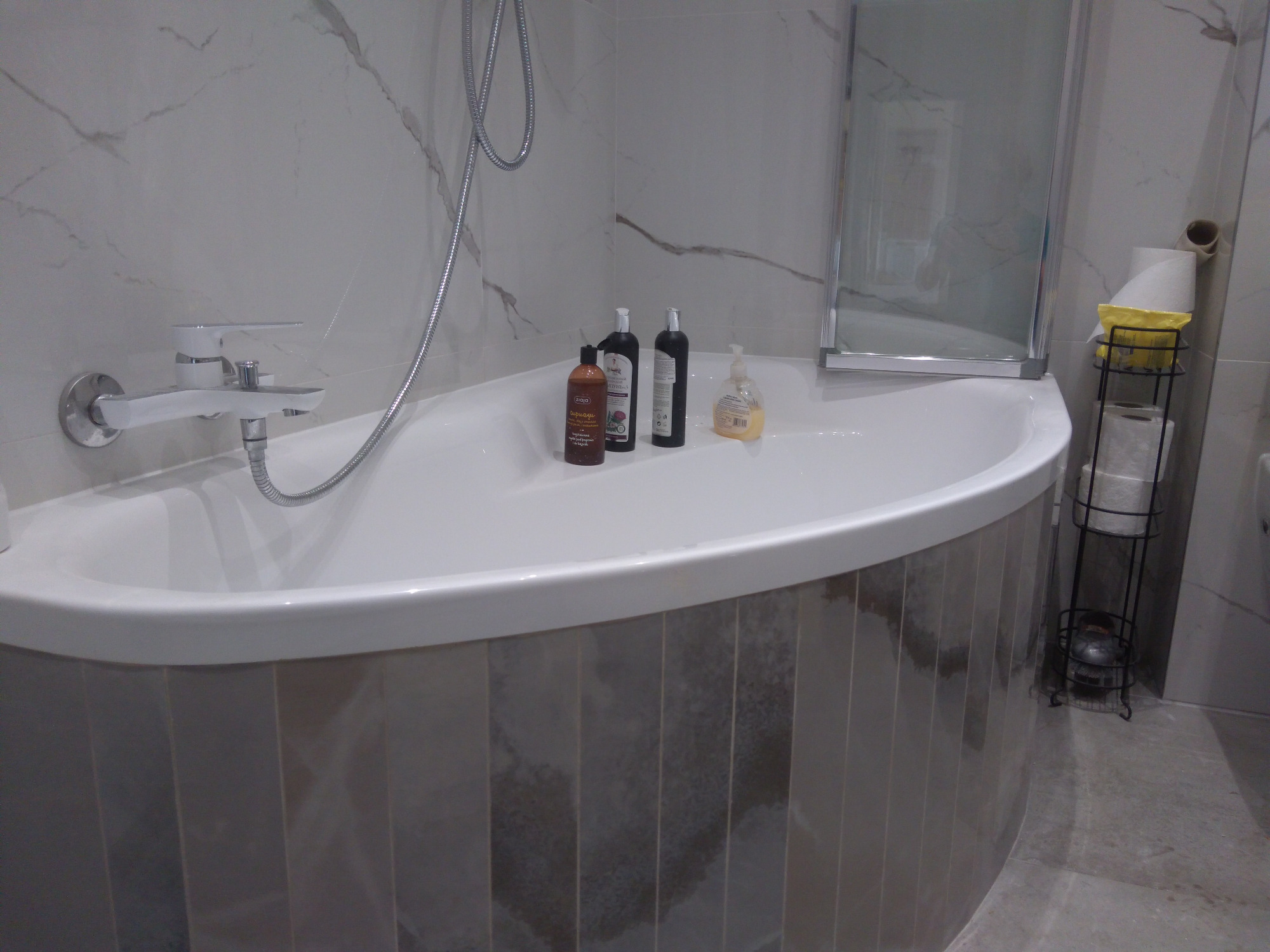 Nowoczesna, owalna wanna obudowana pionowymi płytkami w odcieniach szarości, z chromowaną baterią i słuchawką prysznicową, uzupełniona o butelki z kosmetykami i metalowy stojak na papier toaletowy.