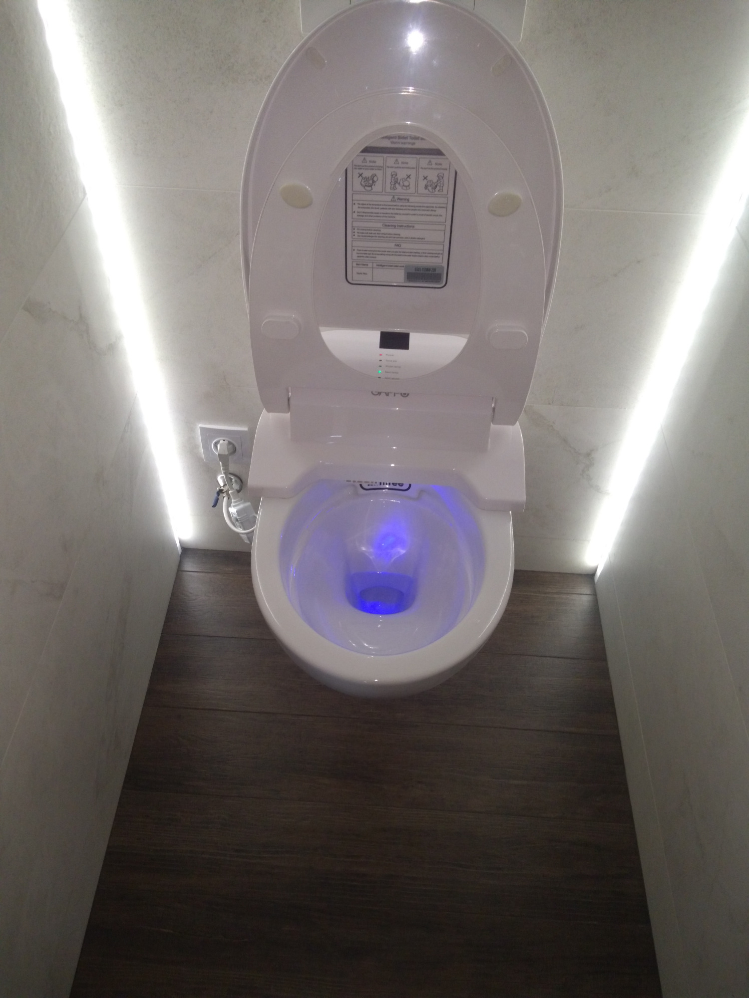 Nowoczesna toaleta z deską myjącą, podświetlana na niebiesko, zamontowana w łazience z płytkami imitującymi marmur i drewnianą podłogą.