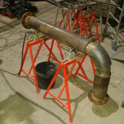 Rusty metalowa rura z kołnierzem, spawana pod kątem 90 stopni, podparta na czerwonych stojakach RIDGID, widoczny spaw na łączeniu segmentów, w tle narzędzia i rusztowania.
