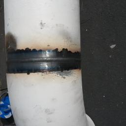 Euro Weld Service - Spoina na rurociągu pomalowanym na biało, widoczne ślady po spawaniu i przypalenia na farbie, obok butelki z wodą i zwinięty kabel.