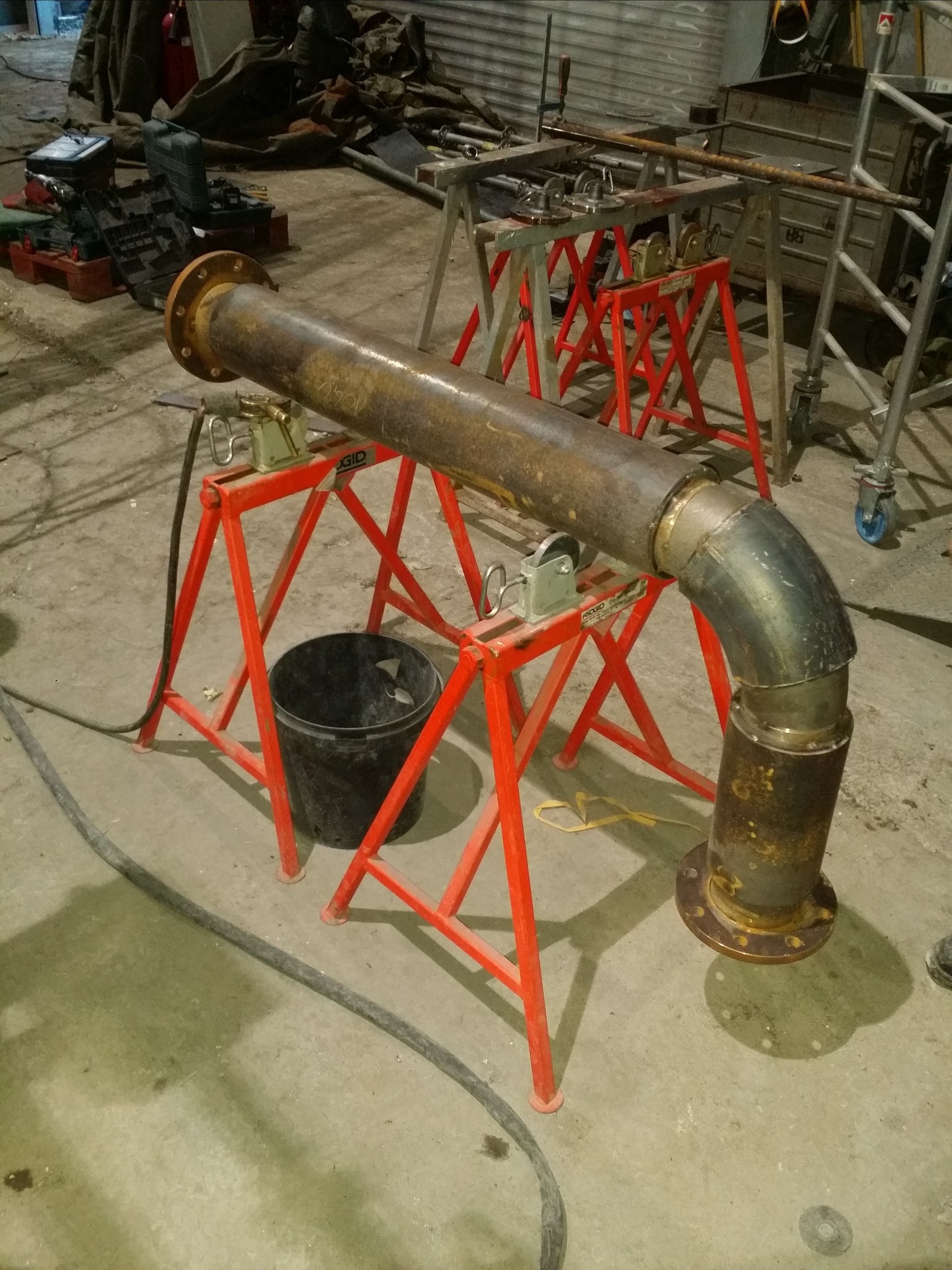 Rusty metalowa rura z kołnierzem, spawana pod kątem 90 stopni, podparta na czerwonych stojakach RIDGID, widoczny spaw na łączeniu segmentów, w tle narzędzia i rusztowania.