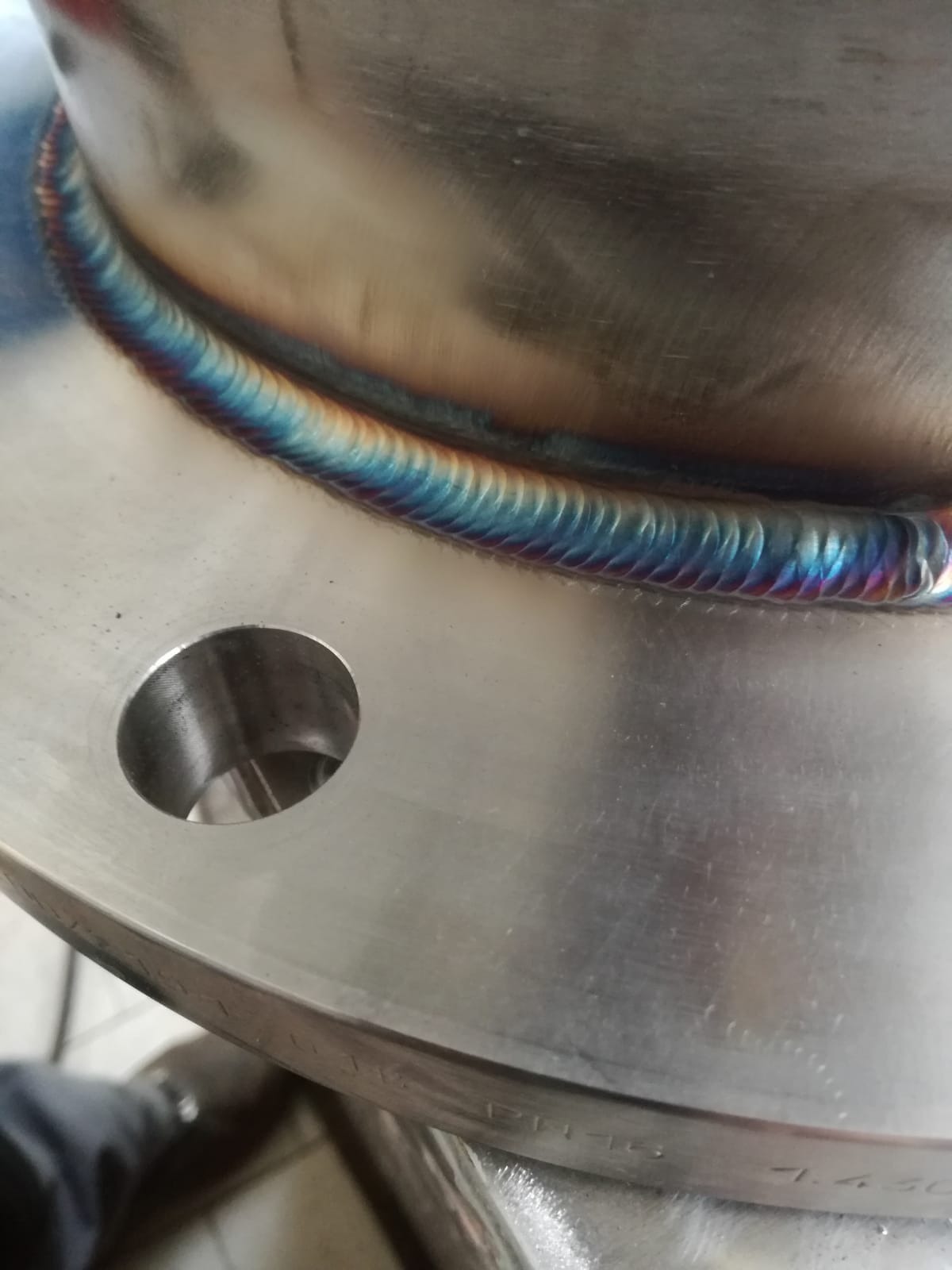 Szczegółowe zbliżenie na spawany kołnierz ze stali nierdzewnej, z widocznymi tęczowymi przebarwieniami spoiny, obok otworu montażowego.