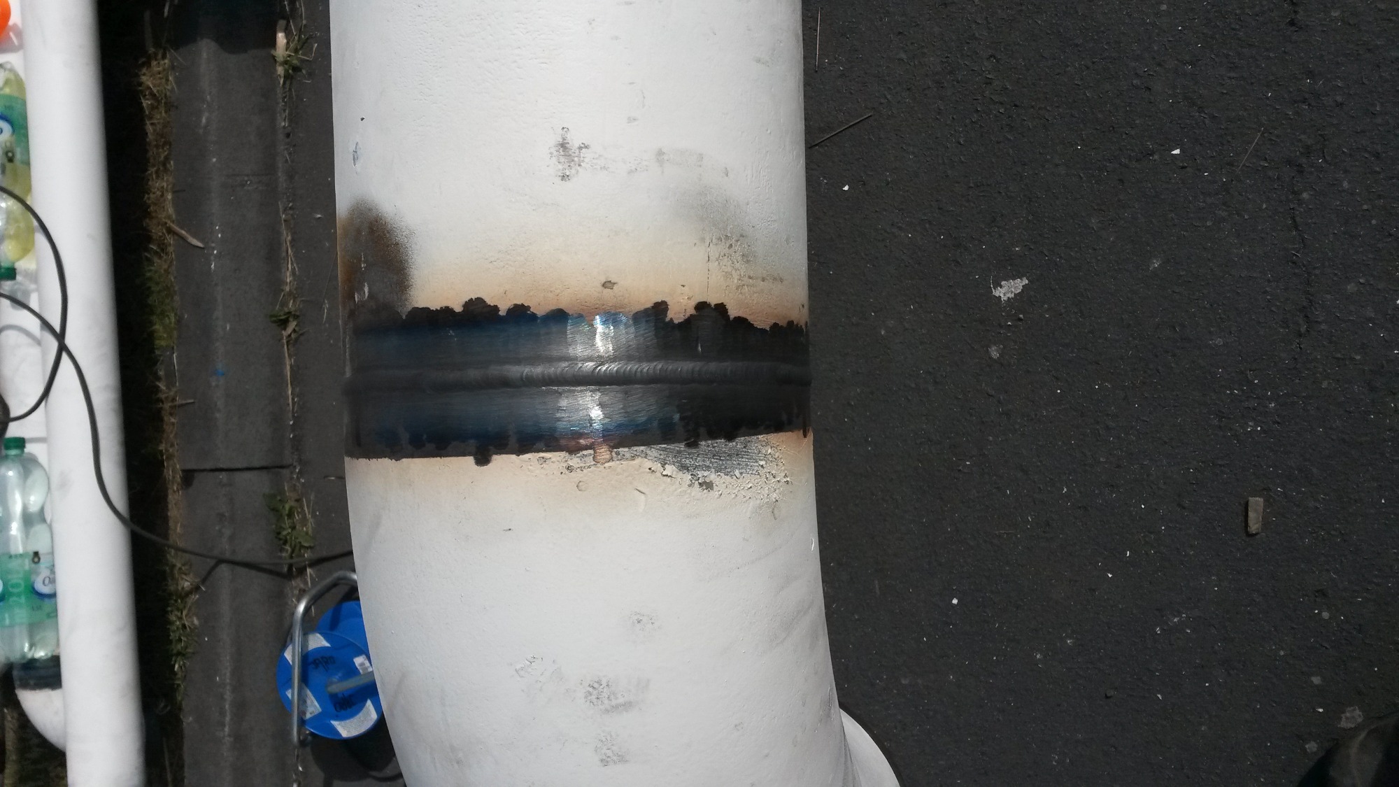Spoina na rurociągu pomalowanym na biało, widoczne ślady po spawaniu i przypalenia na farbie, obok butelki z wodą i zwinięty kabel.
