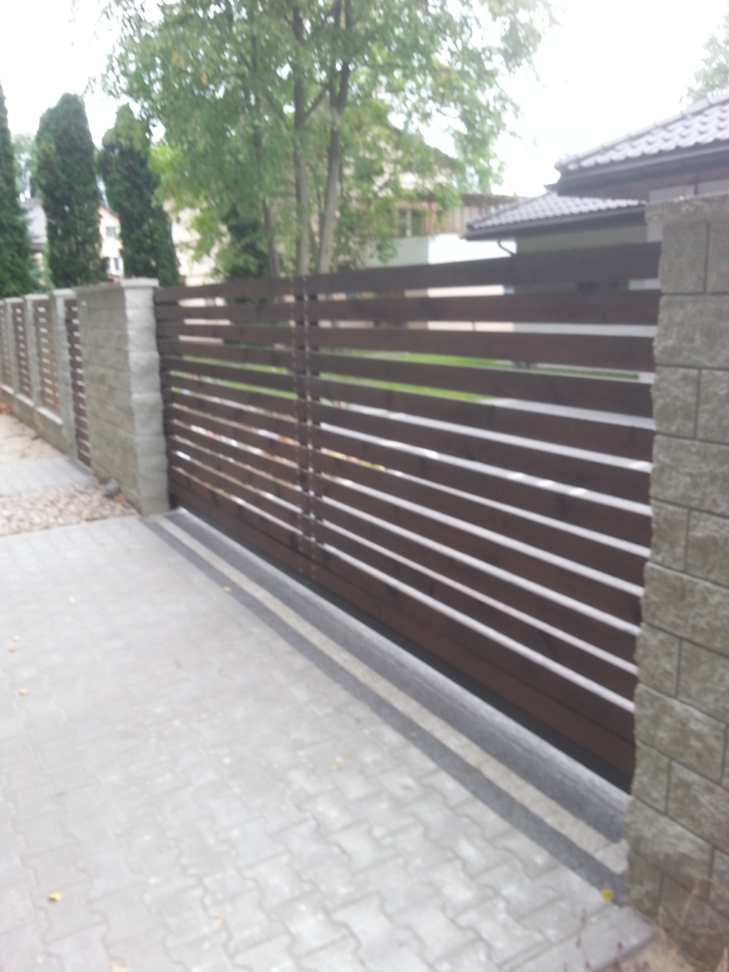 Nowoczesna brama przesuwna z poziomych paneli drewnianych w kolorze brązowym, osadzona między słupkami z szarej kostki, widok z zewnątrz na posesję.