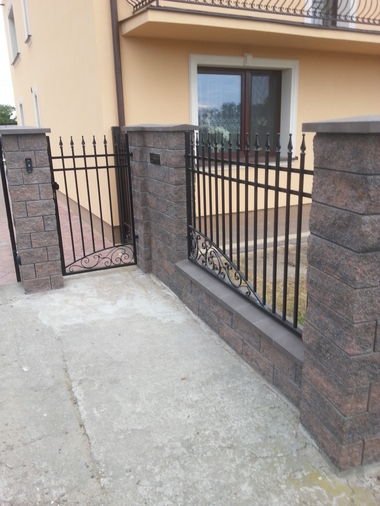 Ciemna, metalowa furtka z dekoracyjnymi elementami, osadzona w murze z brązowych bloczków, na tle elewacji budynku w odcieniach beżu.