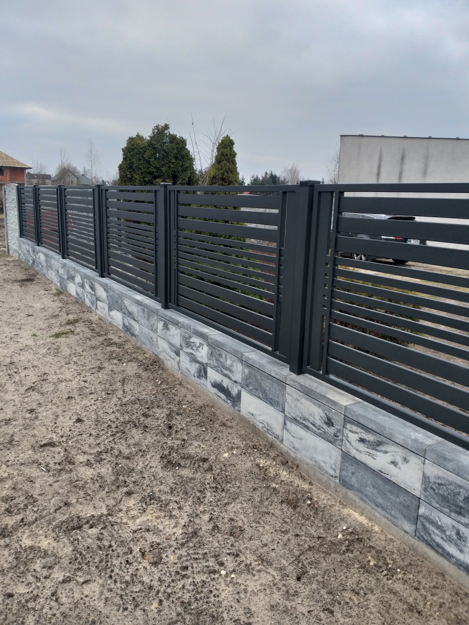 Nowoczesne, poziome, szare przęsła ogrodzeniowe zamontowane na podmurówce z szarych bloczków imitujących kamień, widok z zewnątrz na tle zabudowy jednorodzinnej.