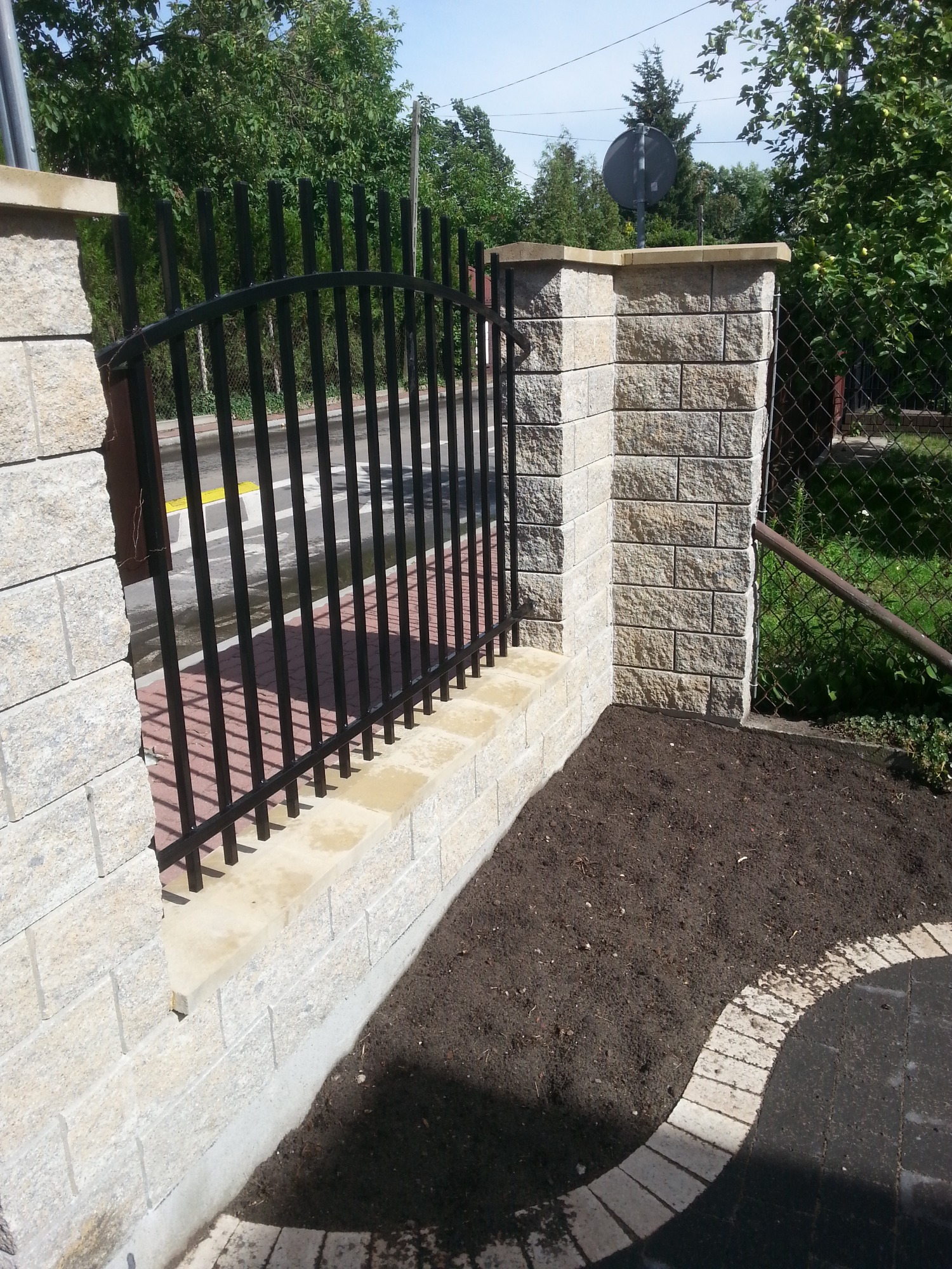 Fragment ogrodzenia z czarnymi prętami i słupkami z jasnego kamienia łupanego, świeżo obsypana ziemia pod ogrodzeniem, widok na ulicę i zieleń w tle.
