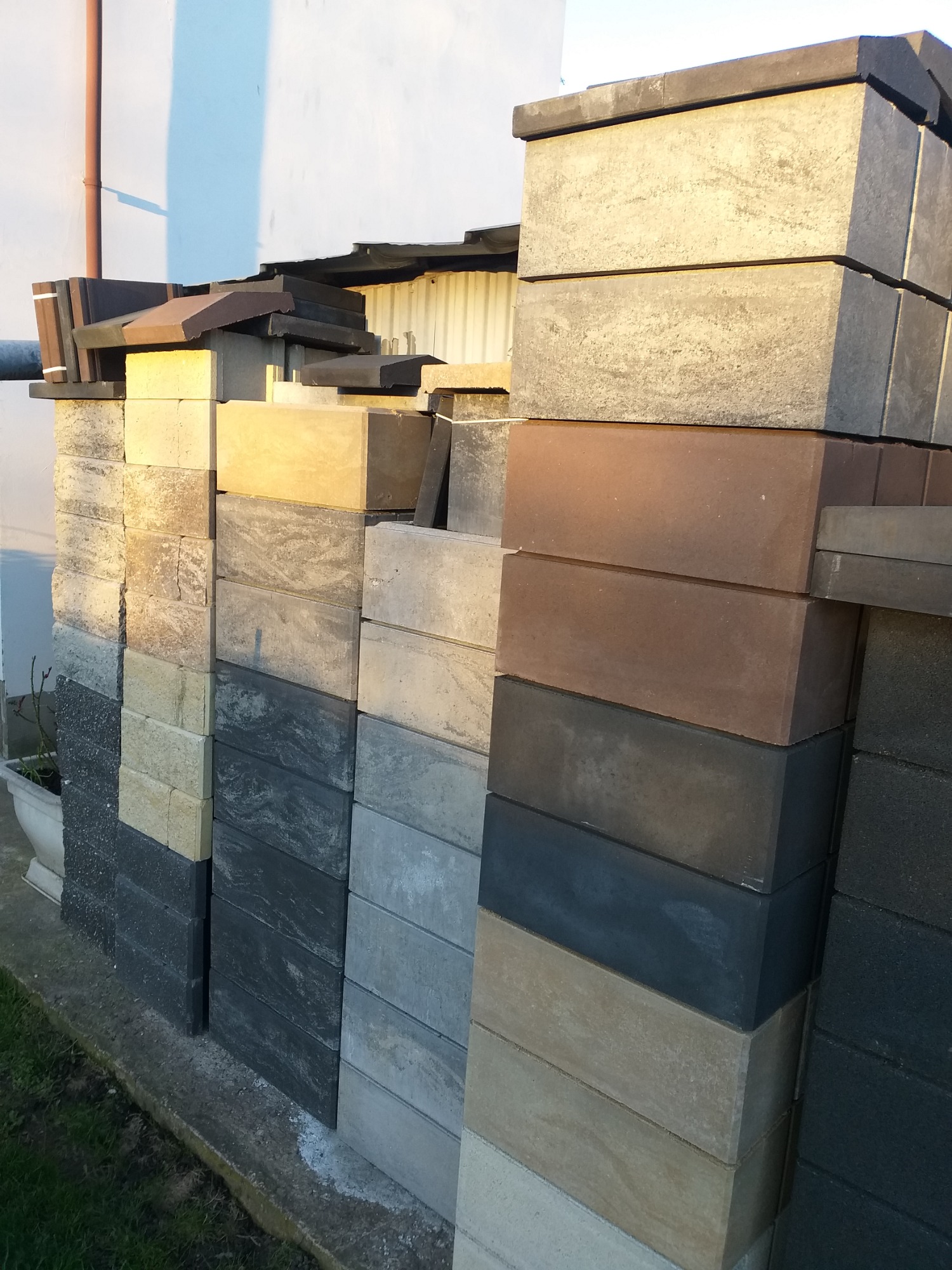 Stosy różnokolorowych bloczków betonowych ułożonych obok białej ściany budynku i zielonego trawnika, widoczne różne odcienie szarości, beżu i brązu, sugerujące materiał do budowy ogrodzenia.