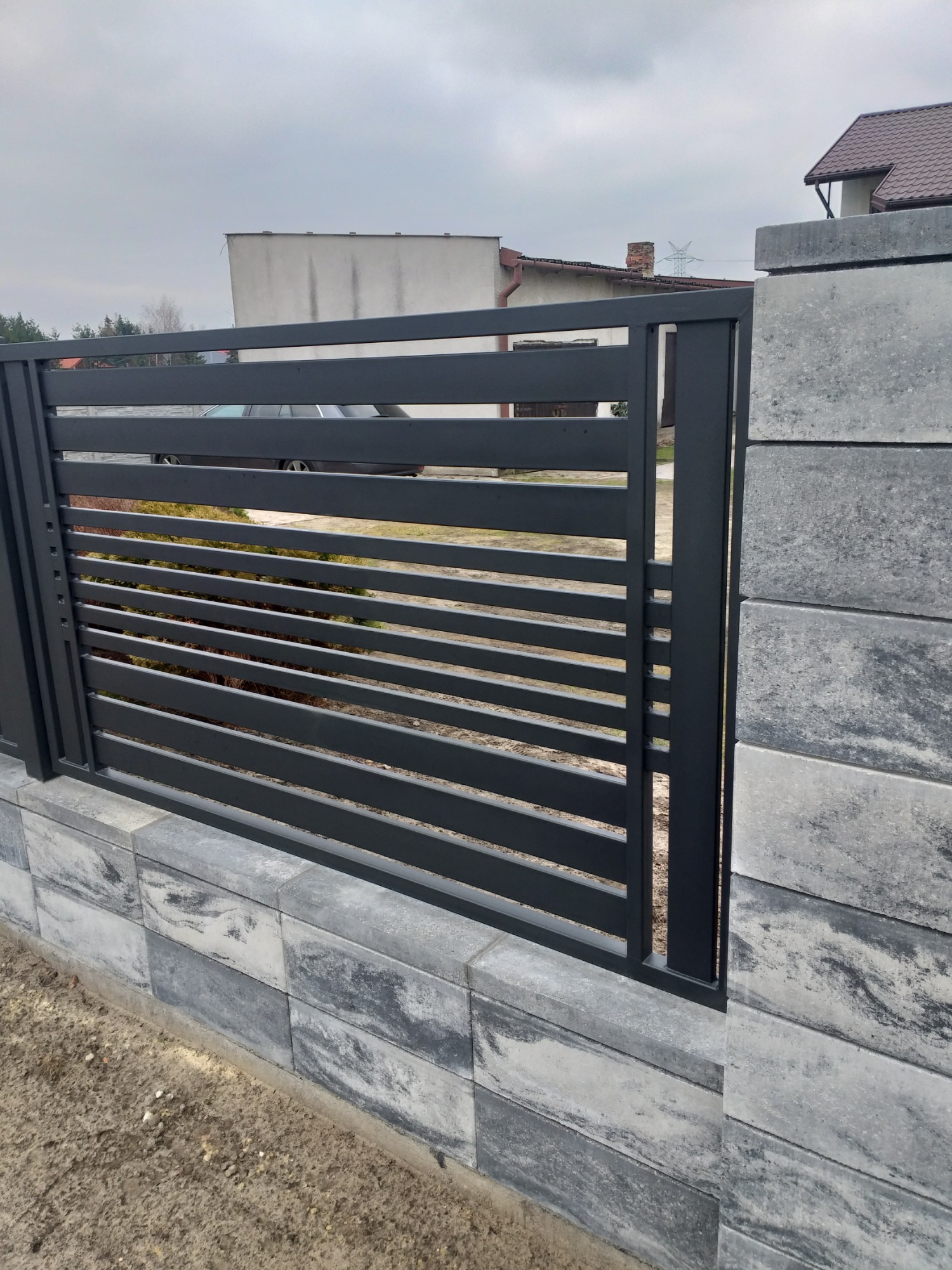 Nowoczesne ogrodzenie panelowe w kolorze grafitowym, zamontowane na podmurówce z szarej kostki betonowej, widok z zewnątrz posesji.