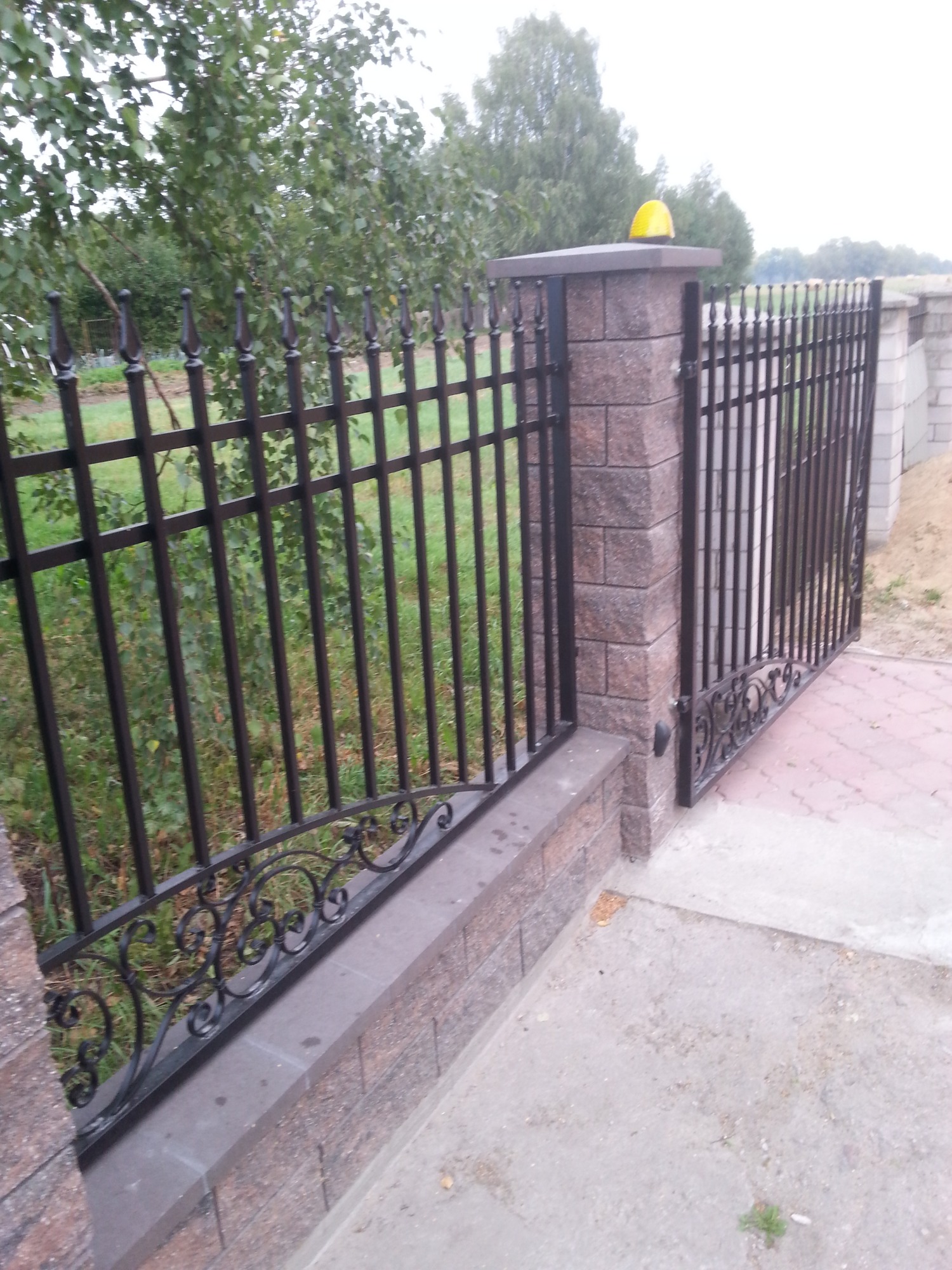 Ciemne, metalowe ogrodzenie panelowe z dekoracyjnymi elementami, osadzone na murowanych słupkach z cegły, z żółtą lampą ostrzegawczą na jednym ze słupków bramy wjazdowej.