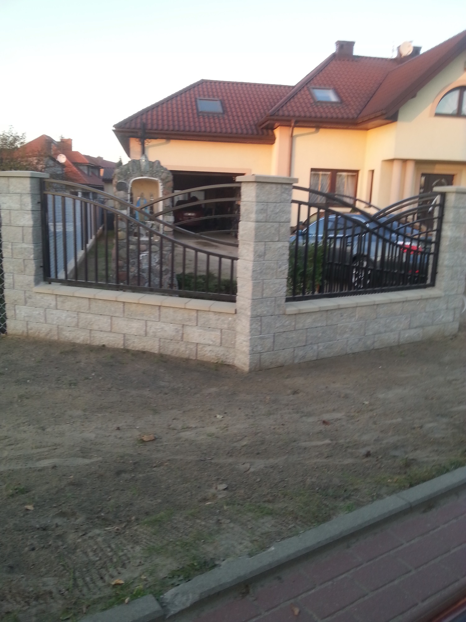 Nowoczesne ogrodzenie z paneli metalowych na podmurówce z szarej kostki, z widoczną kapliczką przydomową i fragmentem domu w tle.