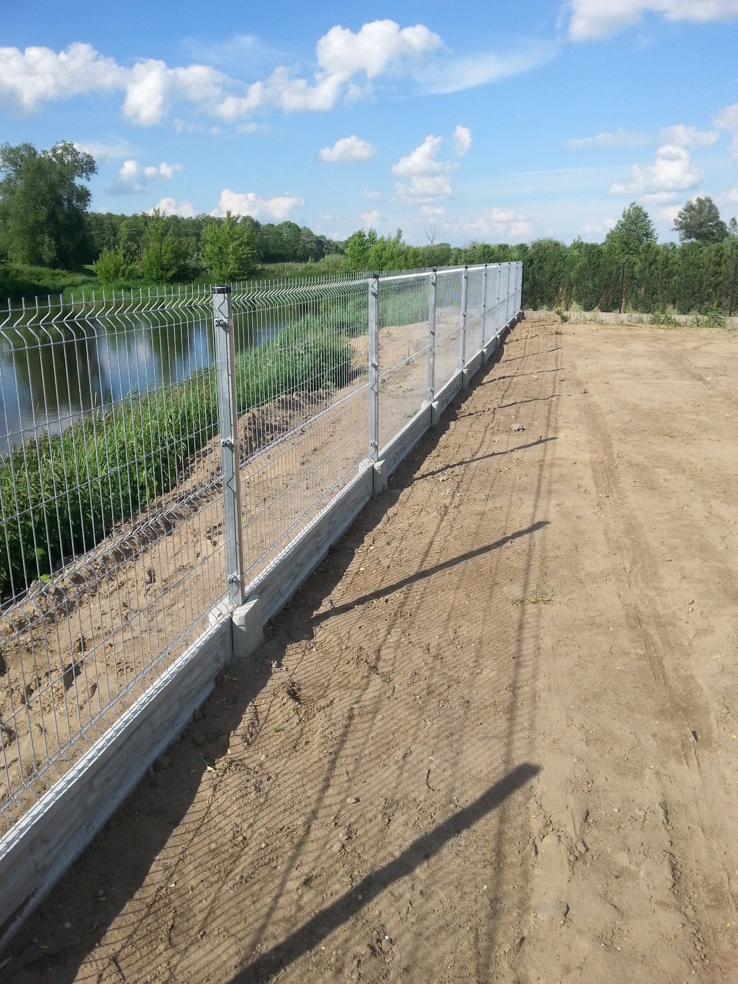 Długi, ocynkowany płot panelowy z podmurówką betonową, biegnący wzdłuż brzegu rzeki z bujną roślinnością, widoczny z perspektywy ziemi z wyraźnymi śladami opon.