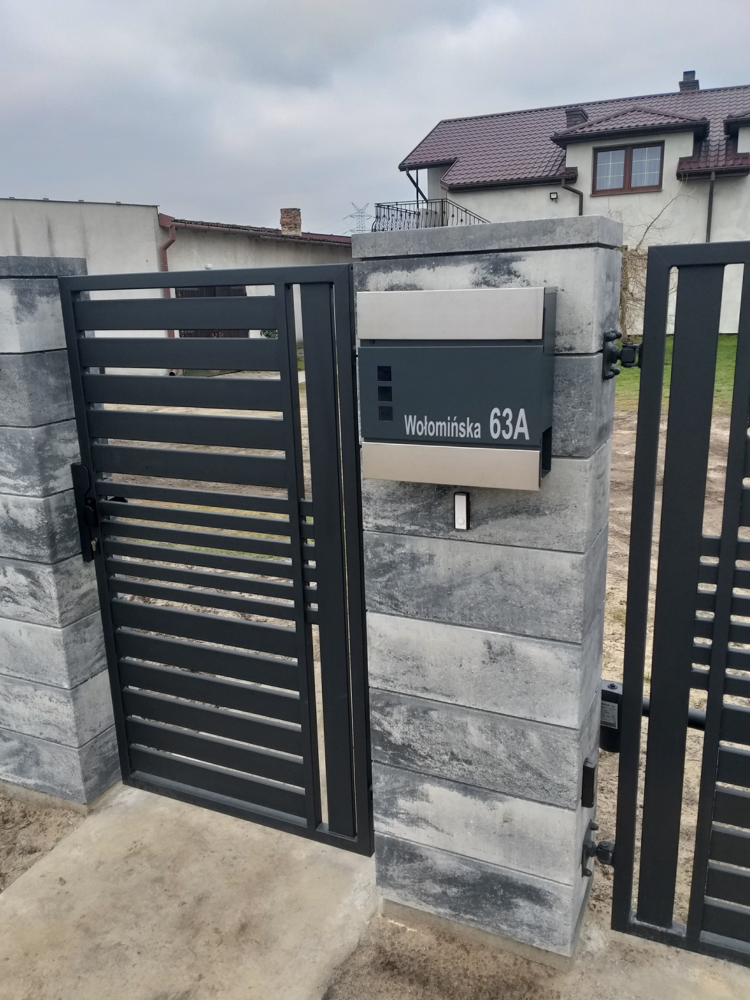 Nowoczesna brama panelowa z poziomymi szczeblami, zamontowana na słupkach z szarej kostki, z przylegającą skrzynką pocztową z numerem 'Wołomińska 63A', na tle domu z brązowym dachem.