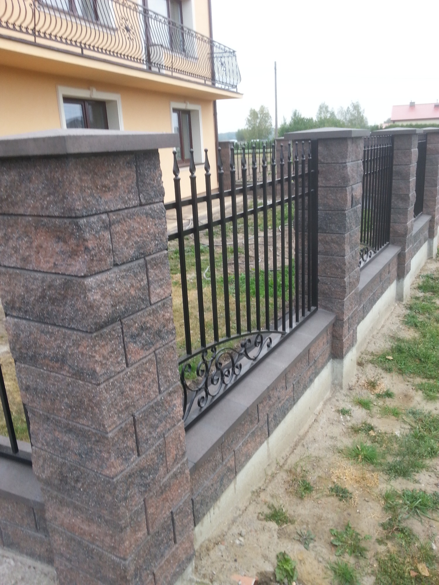 Nowoczesne ogrodzenie z paneli metalowych z ozdobnymi elementami, osadzone na murku z cegły klinkierowej, z widocznym budynkiem w tle.