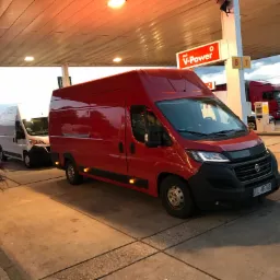 Czerwony dostawczy bus Fiat Ducato zaparkowany na stacji benzynowej Shell, obok biały bus i ciężar&oacute;wka w tle.