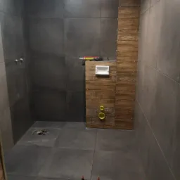 Łazienka w trakcie układania płytek: szare, wielkoformatowe płytki na ścianach i podłodze, imitacja drewna na ścianie z zamontowaną podtynkową spłuczką, widoczne poziomice i krzyżyki dystansowe.