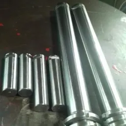 Sześć stalowych sworzni o różnej długości, leżących na brudnym metalowym stole warsztatowym, sfotografowane w sztucznym świetle.